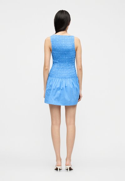 Γυναίκα με μακριά σκούρα μαλλιά, ντυμένη με ένα μίνι φόρεμα χωρίς μανίκια σε ουράνιο γαλάζιο χρώμα με ρελιαστή υφή και λευκά ψηλοτάκουνα mule, στέκεται μπροστά σε μια απλή γκρι φόντο.