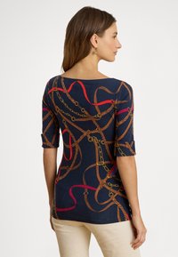 Žena s hnedými vlasmi má na sebe tmavomodrý top so vzorom červených a hnedých reťazí a opaska, béžové nohavice a zlaté náušnice, pozerá sa do strany.