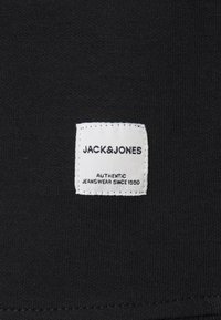 Svart tyg med en vit rektangulär etikett som har "JACK & JONES" och "AUTHENTIC JEANSWEAR SINCE 1990" i svart text.