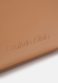 Surface en cuir fauve avec le logo Calvin Klein en relief, montrant une texture fine et une ombre subtile le long du bord inférieur.