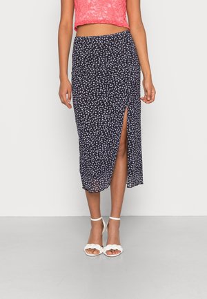 Kvinde iført navyblå blomsterprint midi-nederdel med frontslids, koralfarvet blondetop og hvide hæle med flettet rem på ensfarvet baggrund.