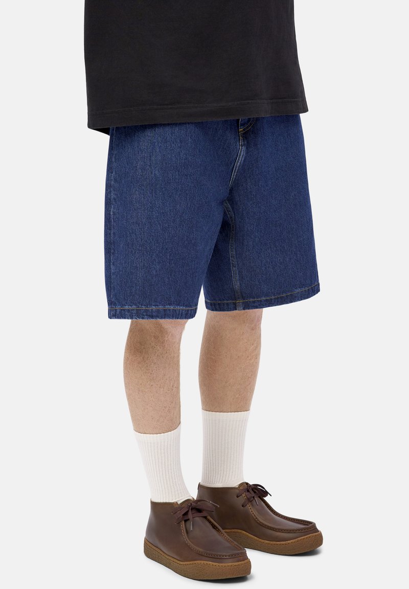 Person wearing mid-length blue denim shorts, white ribbed socks, brown leather moccasin shoes, and a black shirt cropped above the waist.