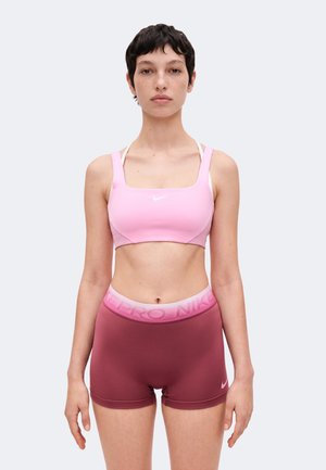 Ung person med kort mørkt hår iført en lyserød sports-bh og mørkerøde Nike Pro shorts står foran en ensfarvet baggrund.