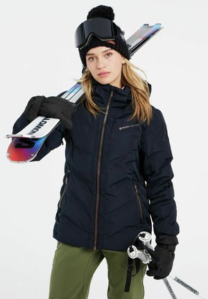 Dunkelblaue Steppjacke mit hohem Kragen und bronzefarbenem Reißverschluss, ergänzt durch eine schwarze Mütze und Goggles. Trägt Skier und weiße Skistöcke.