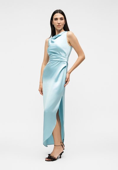 Γυναίκα με μακριά σκουρόχρωμα μαλλιά που φοράει ένα αμάνικο, γαλάζιο σατινέ μίντι φόρεμα με ανοίγματα στο πλάι και μαύρα πέδιλα με λουριά και διακόσμηση από βολάν.