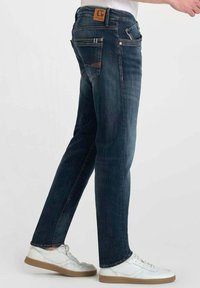 Jeans de mezclilla azul oscuro con un corte recto. Las características incluyen cinco bolsillos, un parche con el logo en la parte trasera y un desgaste mínimo. Llevados con zapatillas blancas.