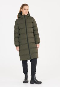 Abrigo acolchado en verde oliva oscuro, con cierre de cremallera frontal, capucha, puños elásticos y dos bolsillos laterales. Combinado con pantalones jogger negros y botas.