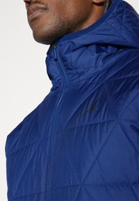 Mornarska quiltana jakna z zadrgo spredaj in kapuco. Ima subtilen črn logotip na levem prsnem delu in rebraste robove ovratnika.