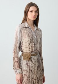 Young woman wearing long-sleeve, buttoned beige snake print dress with matching belt bag, standing against a plain light background.