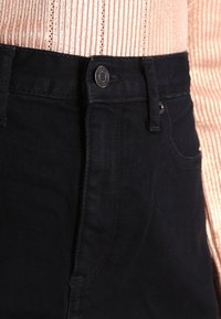 Taille de jean en denim noir avec un bouton métallique attaché sur un pull en maille côtelée rose clair.