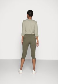 Henkilö seisoo selkä kameraan, päällään vaalea harmaa pitkähihaine paita, oliivinvihreät capri-housut ja valkoiset lenkkarit yksinkertaista taustaa vasten.