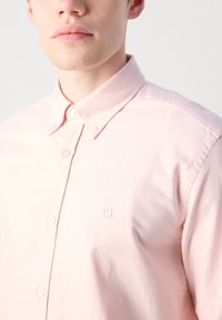 Ungdom som har på seg en lys rosa skjorte med knapper og en liten brodert logo på brystet, avkortet ved nakken og skuldrene.