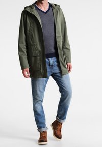 Olivgrön vattentät jacka med huva, framfickor, bärs över en marinblå tröja, kombinerad med ljusblå jeans och bruna stövlar.