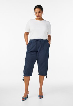 Kvinde iført hvid kortærmet top, marineblå cropped bukser med snøre og lommer, samt blå åbne høje hæle, stående med hænderne i lommerne.