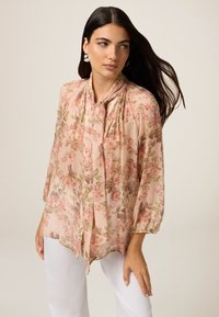 Blusa floreale in tessuto rosa chiaro trasparente con colletto a fiocco, motivi floreali in verde e rosa scuro, e maniche a 3/4 a sbuffo.