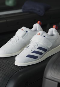 White athletic shoes with a textured surface, featuring a blue accent stripe design and red heel loop, secured with a strap and laces.