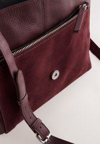 Bourgondische tas met een mix van getextureerd leer en suède, voorzien van een ritssluiting, drukknopsluiting en een verstelbare riem.