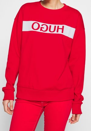 Osoba ubrana w czerwoną bluzę i spodnie, bluza ma biały pasek z odwróconym logo HUGO na wysokości klatki piersiowej.