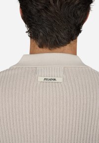 Lysbeige ribbet polo skjorte med en kontrasterende krave, der har et lille mærke med teksten "PEGADOR" på nakken. Tekstureret stof.