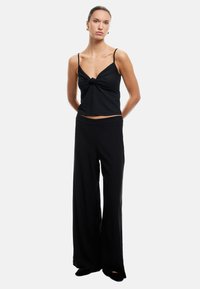 Top de tanque negro de tricot con un detalle de nudo en la parte delantera, combinado con pantalones negros de corte holgado. El material parece suave con una textura sutil.