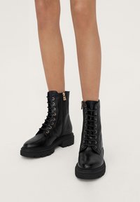 Botas de cuero negro hasta el tobillo con ojales dorados, cordones y cremallera lateral. Suelas gruesas negras con textura. Acabado liso, altura media hasta el tobillo.
