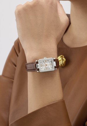 Quadratische silberne Uhr mit braunem Lederarmband am Handgelenk, getragen von einer Person in braunem Langarmshirt mit goldener Zierknopfleiste.