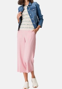 Modelka v džínové bundě přes pruhované zeleno-růžové tričko, růžových širokých zkrácených kalhotách a bílých teniskách, pózuje s rukama v kapsách.