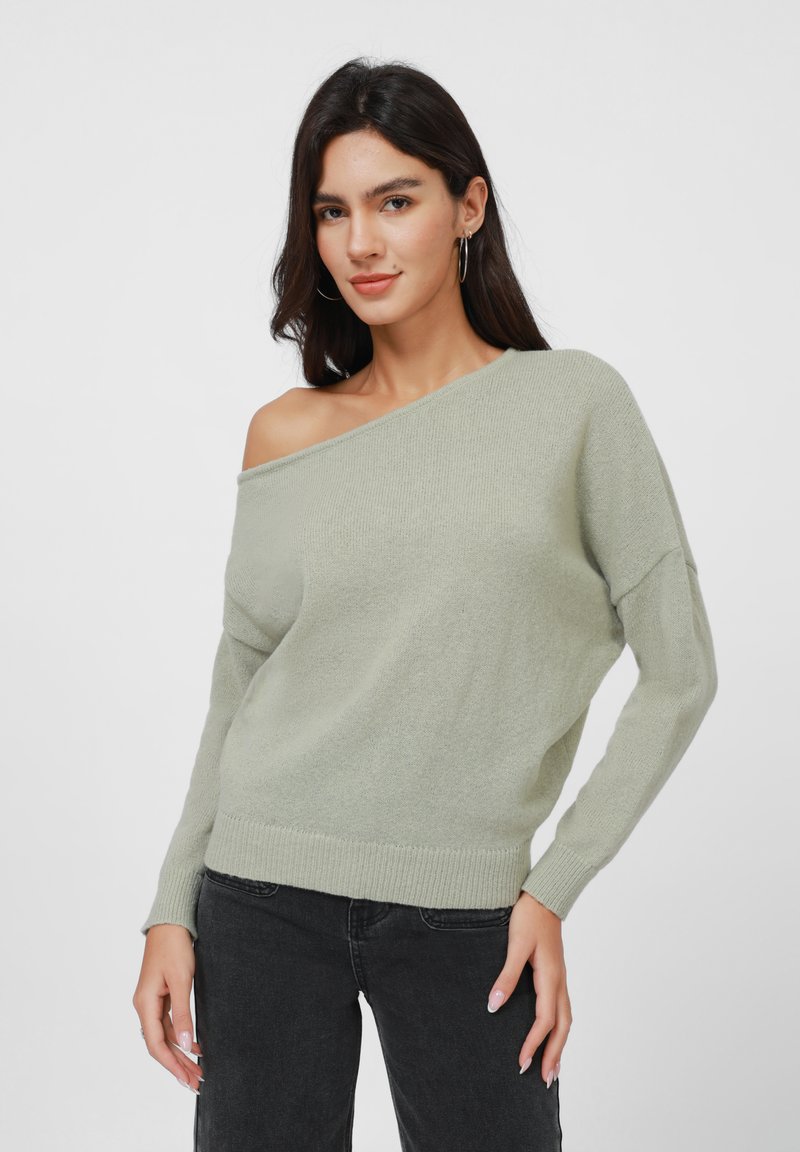 Lys grønn oversized genser med avskåret design, myk tekstur, ribbede mansjetter og hem; sammen med svarte jeans.
