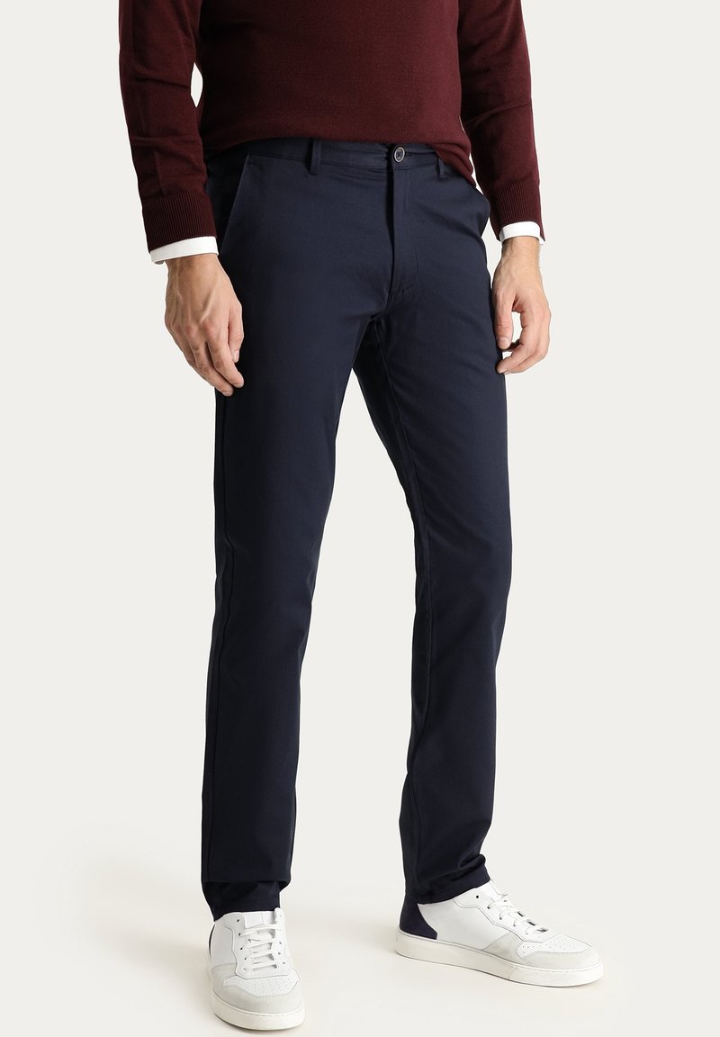 Homme portant un pantalon slim bleu marine foncé, des baskets blanches et une chemise à manches longues bordeaux avec des poignets blancs, debout devant un fond uni.