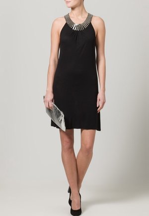 Musta hihaton mekko, jossa on laskostettu hopeahelmikoristeinen pääntie. Mekko on vartaloa myötäilevä ja hieman liehuva, yhdistetty metallinhohtoiseen pikkulaukkuun ja mustiin korkokenkiin.