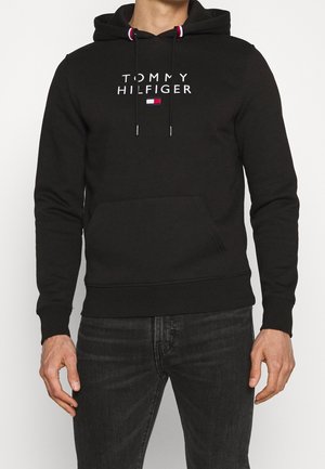 Mężczyzna ubrany w czarną bluzę z kapturem Tommy Hilfiger z przednią kieszenią i tekstowym logo, w połączeniu z ciemnoszarymi dżinsami, stojący na jednolitym tle.