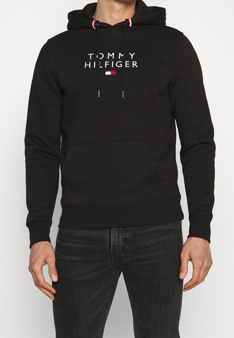 Homme portant un sweat à capuche noir Tommy Hilfiger avec poche avant et texte logo, associé à un jean gris foncé, debout devant un fond uni.