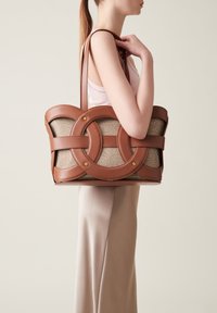 Brown leather tote bag featuring interlinked circular cutouts and a textured beige fabric body. Two shoulder straps and gold hardware accents.