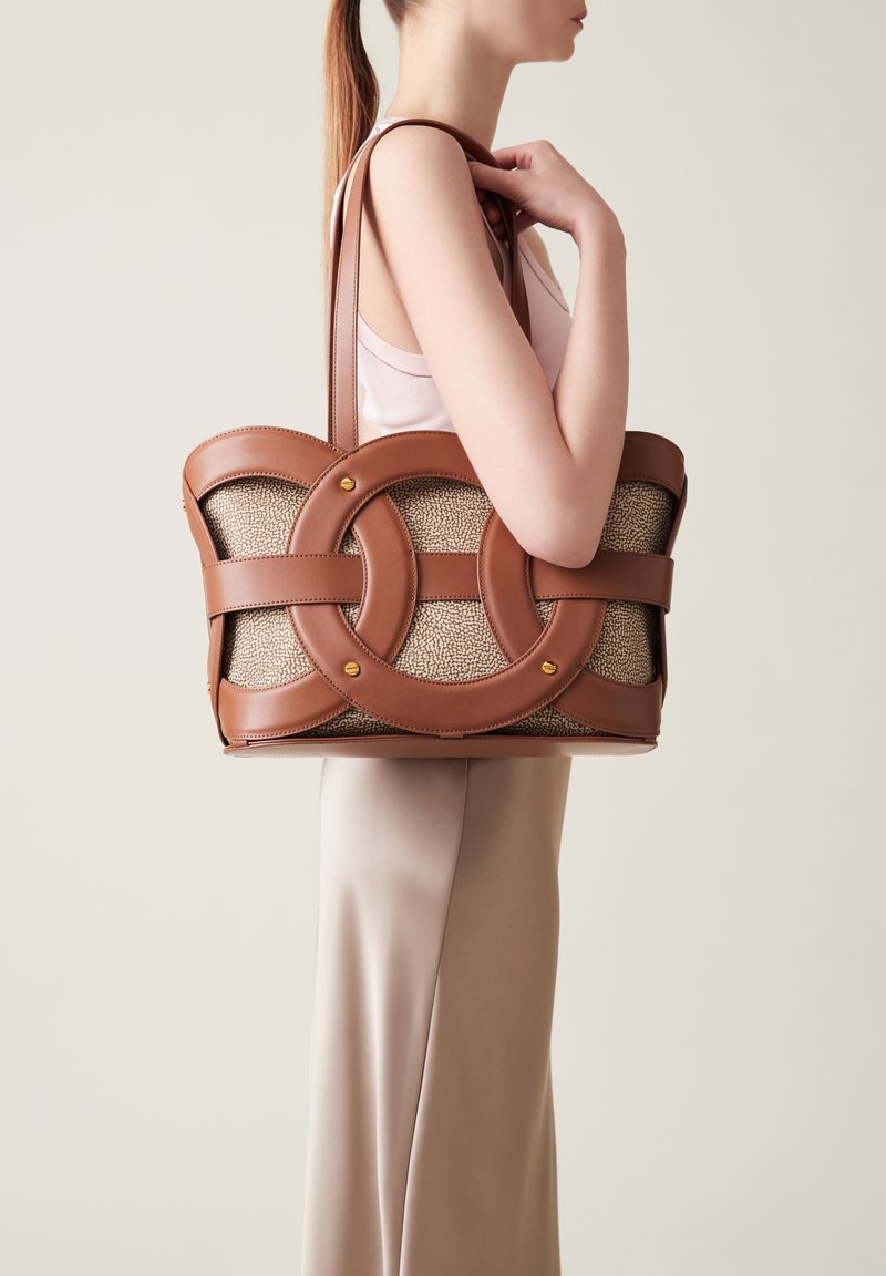 Brown leather tote bag featuring interlinked circular cutouts and a textured beige fabric body. Two shoulder straps and gold hardware accents.