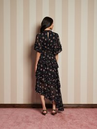 Vestido preto de chiffon com padrão floral, camadas rizadas e bainha assimétrica. Detalhe de abertura nas costas visível. Combinado com saltos de estampa de leopardo.