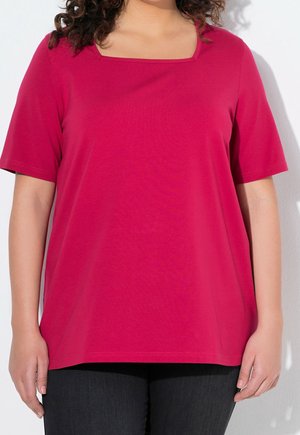 Vrouw draagt een felroze top met korte mouwen en een vierkante halslijn en donkere broek, staand tegen een witte achtergrond.