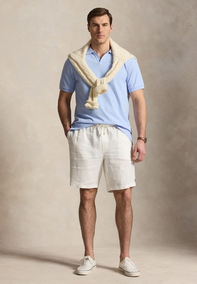 Άνδρας με ανοιχτό μπλε πόλο, λευκά σορτς με κορδόνι, λευκά αθλητικά παπούτσια και εκρού πουλόβερ περασμένο πάνω από τους ώμους σε μονόχρωμο φόντο.