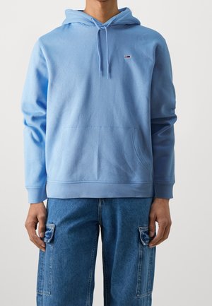 Mężczyzna w jasnoniebieskiej bluzie z kapturem i kieszenią z przodu oraz niebieskich spodniach typu cargo na gładkim tle.