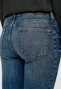 Jeans en denim bleu avec une taille mi-haute, coupe slim et poches arrière avec une texture légèrement décolorée. Couture dans une couleur contrastante.