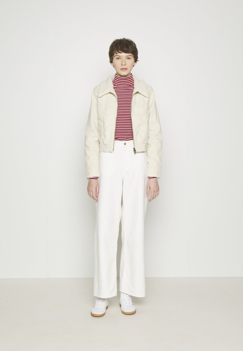 Personne portant une veste crème avec un col en fausse fourrure, un pull à col roulé rouge et blanc rayé, un pantalon large blanc et des baskets blanches, se tenant devant un mur uni.