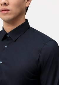 Navy Hemdbluse aus glattem Stoff, mit klassischem Kragen, blauen Knöpfen und taillierter Passform. Nahaufnahme des Schulter- und Kragenbereichs.