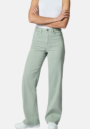 Personne portant un pantalon large taille haute vert clair et des baskets blanches, debout bras croisés devant un fond uni.