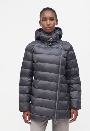 Femme portant une doudoune noire matelassée avec une fermeture éclair asymétrique et un col haut sur un fond blanc uni.