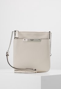 Bolso cruzado beige de Guess con placa de logo metálica plateada, cierre con cremallera y correa ajustable con detalle de cadena sobre una superficie blanca.