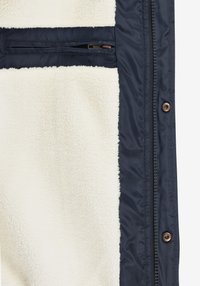 Vonkajšia bunda s podšívkou z flísu, námornícka modrá vonkajšia strana a krémová vnútorná časť. Má zipsový náprsné vrecko a bronzové kovanie.