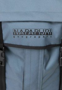 Mochila de tela azul con correas negras, textura suave y un logotipo negro destacado con "NAPAPIJRI" y "geographic" impresos.