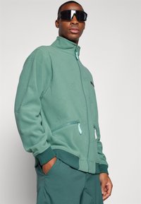 Grønn fleece zipjakke med høy krage, med glidelås-lommer og pregete mønstre. Brukt sammen med matchende grønne bukser og solbriller.
