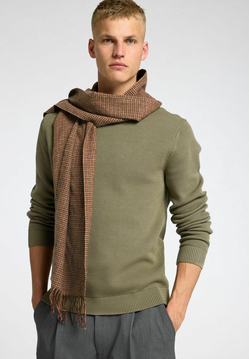 Sciarpa a scacchi marrone drappeggiata su un maglione verde oliva con texture. Il maglione ha polsini e orlo a coste, mentre la sciarpa presenta bordi frangiati.