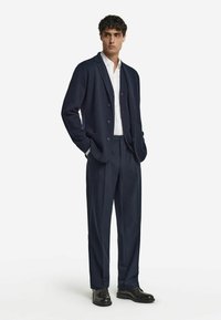 Homme standing avec les mains dans les poches, portant un blazer bleu marine, un pantalon assorti, une chemise blanche et des chaussures de ville noires sur un fond blanc.