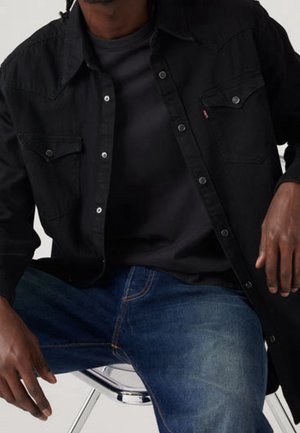 Homme portant une chemise noire à boutons sur un t-shirt noir et un jean bleu, assis sur une chaise en métal avec une main posée sur la cuisse.